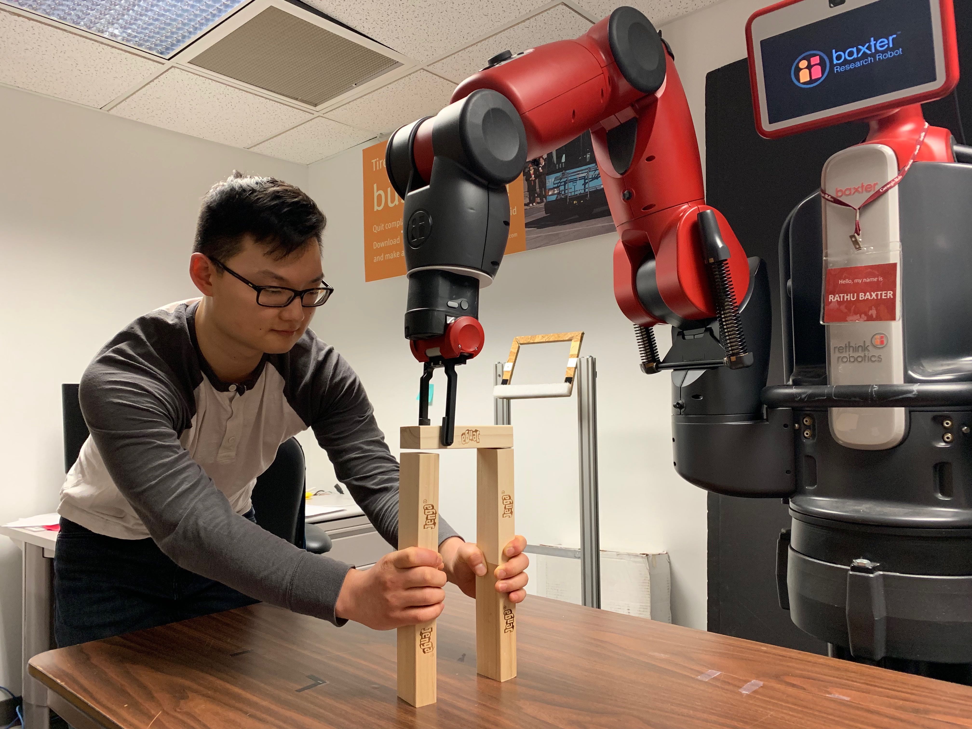 A Baxter humanoid robot uses its gripper on its arm to stack a Jenga block on top of the tower that a human is constructing.